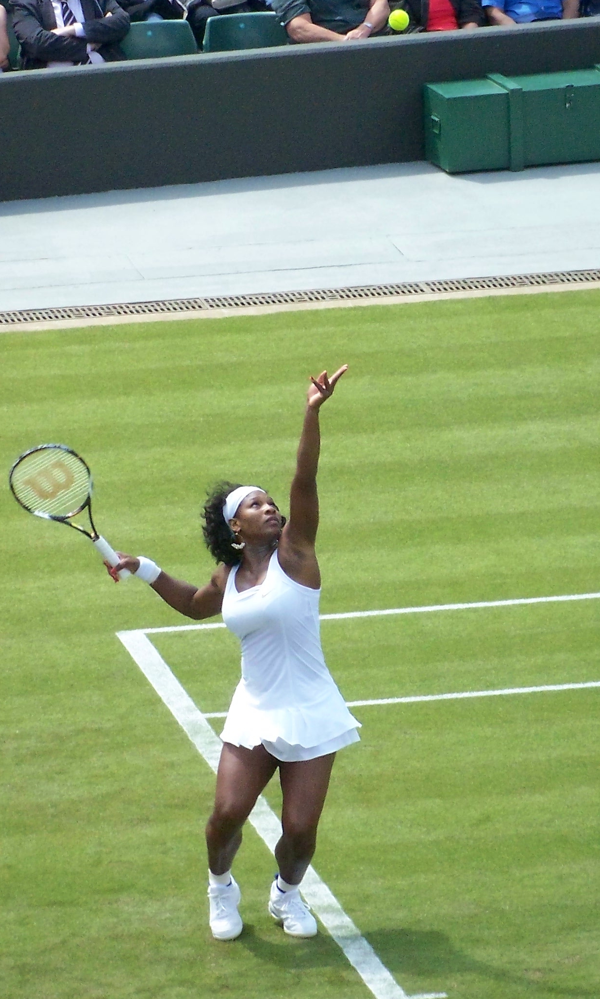Serena Williams in a white tennis dress preparing to serve on a grass court.